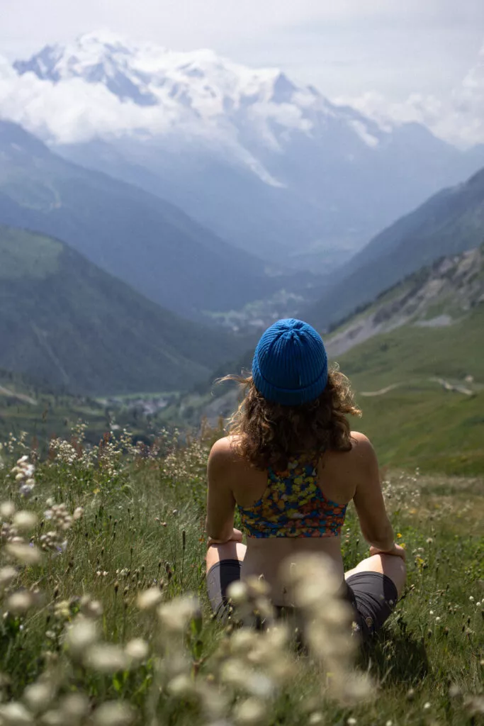 Osoba v modré čepici a barevném topu ze 100% merina vlny sedí se zkříženýma nohama na horské louce, s výhledem na zasněžené štíty pod zataženou oblohou.
