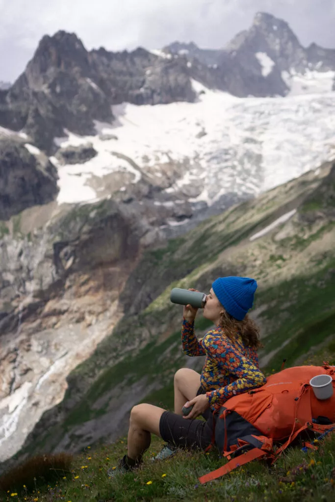 Osoba v modré čepici a barevné bundě z wełny merino 100% sedí na trávě u oranžového batohu, pije z lahve. V pozadí zasněžené hory.