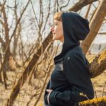 Žena v dámské Merino mikině na zip s kapucí - černá (250 g) z wełny merino 100 % stojí venku ve slunném, bezlistém lese.