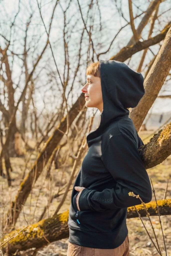 Žena v dámské Merino mikině na zip s kapucí - černá (250 g) z wełny merino 100 % stojí venku ve slunném, bezlistém lese.