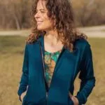 Usmívající se žena s kudrnatými vlasy stojí venku na trávě, má na sobě Merino mikinu na zip s kapucí dámskou - petrol (250 g) z wełny merino 100 %, ruce v kapsách přes barevnou košili. V pozadí jsou vidět bezlisté stromy.