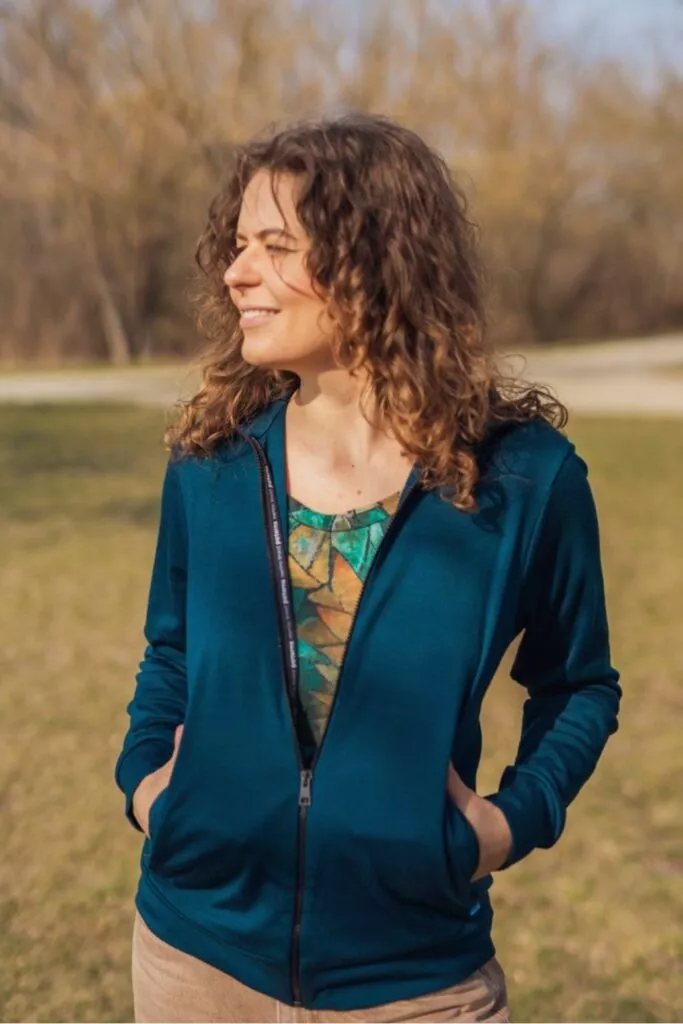 Usmívající se žena s kudrnatými vlasy stojí venku na trávě, má na sobě Merino mikinu na zip s kapucí dámskou - petrol (250 g) z wełny merino 100 %, ruce v kapsách přes barevnou košili. V pozadí jsou vidět bezlisté stromy.