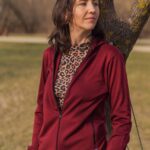 Žena stojí venku před stromem v mikině Merino na zip s kapucí dámské - bordo (250 g) z wełny merino 100%, pod ní leopardí top, béžové kalhoty, dívá se stranou s neutrálním výrazem.