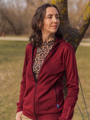Žena stojí venku před stromem v mikině Merino na zip s kapucí dámské - bordo (250 g) z wełny merino 100%, pod ní leopardí top, béžové kalhoty, dívá se stranou s neutrálním výrazem.