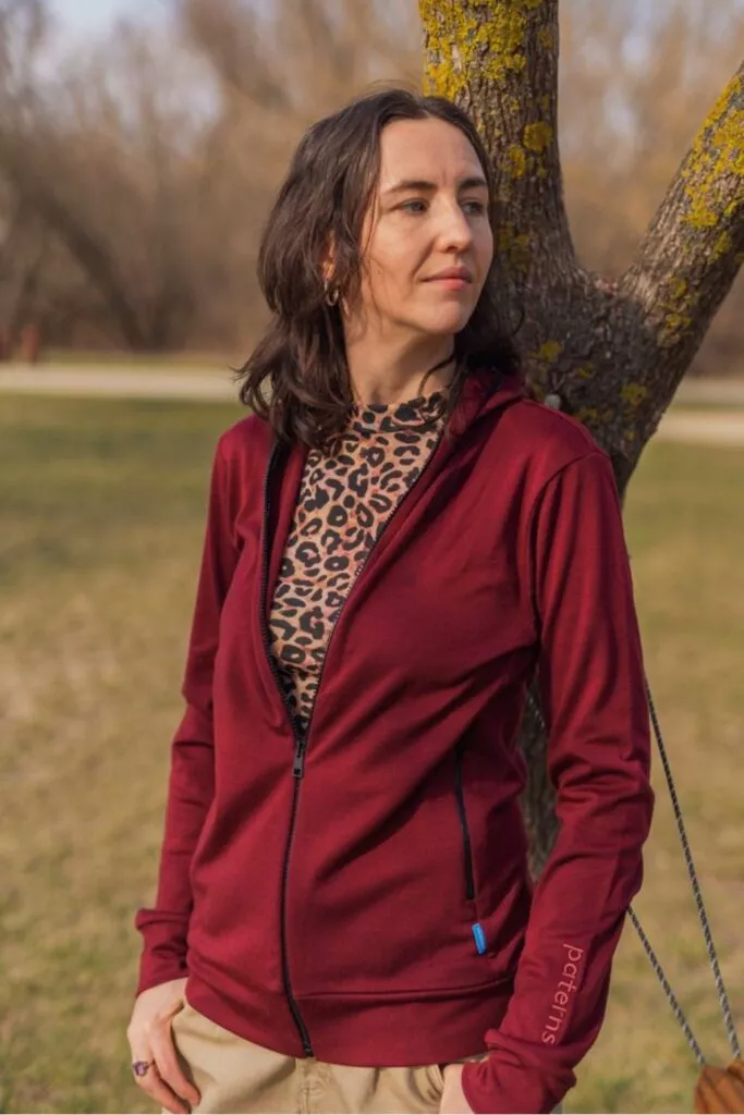 Žena stojí venku před stromem v mikině Merino na zip s kapucí dámské - bordo (250 g) z wełny merino 100%, pod ní leopardí top, béžové kalhoty, dívá se stranou s neutrálním výrazem.