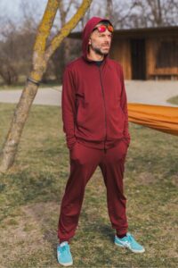 Muž venku s rukama v kapsách má na sobě Merino mikinu na zip s kapucí pánskou - bordo (250 g) ze 100% merino vlny. Vzadu stromy, tráva a dřevěná stavba.