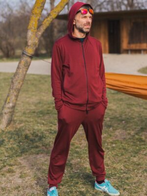 Muž venku s rukama v kapsách má na sobě Merino mikinu na zip s kapucí pánskou - bordo (250 g) ze 100% merino vlny. Vzadu stromy, tráva a dřevěná stavba.