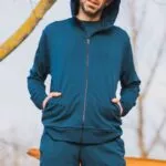 Muž stojí venku s rukama v kapsách, má na sobě Merino mikinu na zip s kapucí pánskou - petrol (250 g, z wełny merino 100%) a červené brýle. V pozadí jsou holé stromy a oranžový předmět.