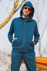 Muž stojí venku s rukama v kapsách, má na sobě Merino mikinu na zip s kapucí pánskou - petrol (250 g, z wełny merino 100%) a červené brýle. V pozadí jsou holé stromy a oranžový předmět.