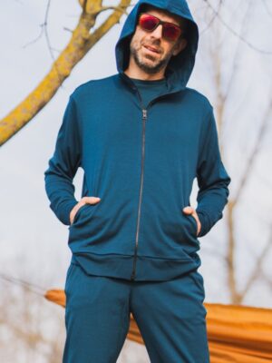 Muž stojí venku s rukama v kapsách, má na sobě Merino mikinu na zip s kapucí pánskou - petrol (250 g, z wełny merino 100%) a červené brýle. V pozadí jsou holé stromy a oranžový předmět.