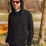 Muž v mikině Merino na zip s kapucí pánskou - černá (250 g) z vlny merino 100 % stojí venku s rukama v kapsách, nasazenými červenými slunečními brýlemi a stromy s rozmazaným pozadím parku za zády.