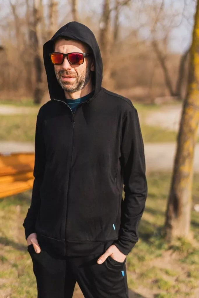 Muž v mikině Merino na zip s kapucí pánskou - černá (250 g) z vlny merino 100 % stojí venku s rukama v kapsách, nasazenými červenými slunečními brýlemi a stromy s rozmazaným pozadím parku za zády.