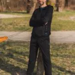 Muž má na sobě Merino mikinu na zip s kapucí pánskou - černá (250 g) ze 100% merino vlny, stojící venku s překříženýma rukama, opřený o strom. Usmívá se v červených slunečních brýlích, modrých teniskách a černých teplácích, za ním zelená tráva a cesty.