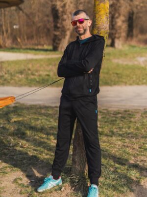 Muž má na sobě Merino mikinu na zip s kapucí pánskou - černá (250 g) ze 100% merino vlny, stojící venku s překříženýma rukama, opřený o strom. Usmívá se v červených slunečních brýlích, modrých teniskách a černých teplácích, za ním zelená tráva a cesty.