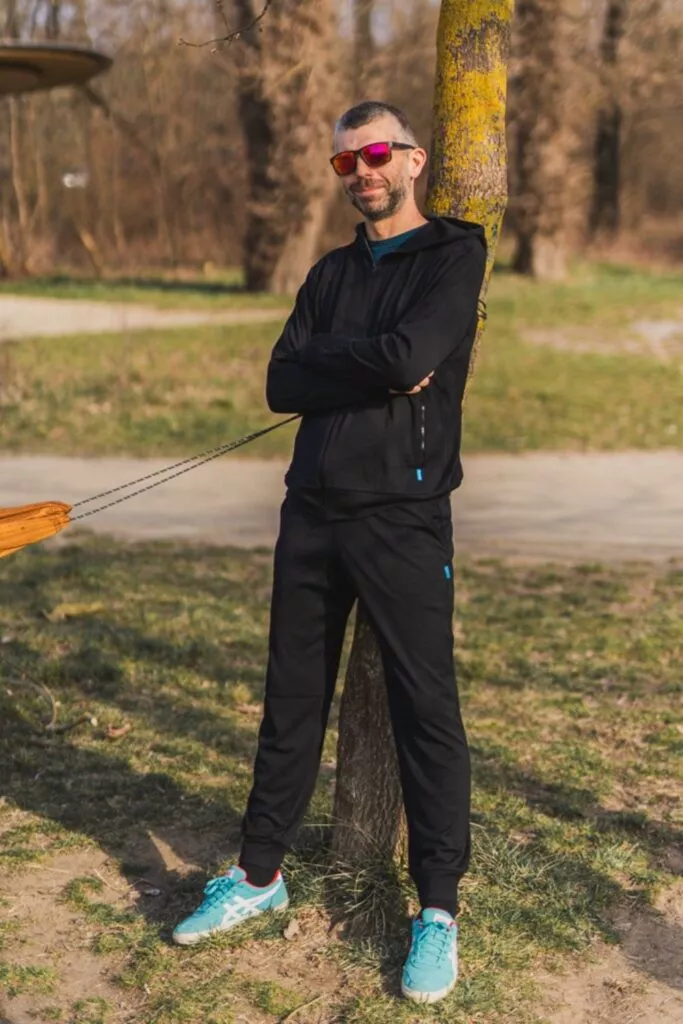 Muž má na sobě Merino mikinu na zip s kapucí pánskou - černá (250 g) ze 100% merino vlny, stojící venku s překříženýma rukama, opřený o strom. Usmívá se v červených slunečních brýlích, modrých teniskách a černých teplácích, za ním zelená tráva a cesty.