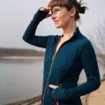 Žena v Merino mikině sportovní dámské - petrol (250 g) z wełny merino 100% stojí venku u vody, stíní si oči a usmívá se.