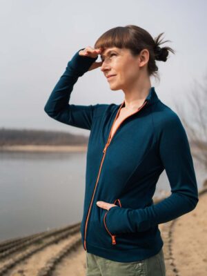 Žena v Merino mikině sportovní dámské - petrol (250 g) z wełny merino 100% stojí venku u vody, stíní si oči a usmívá se.