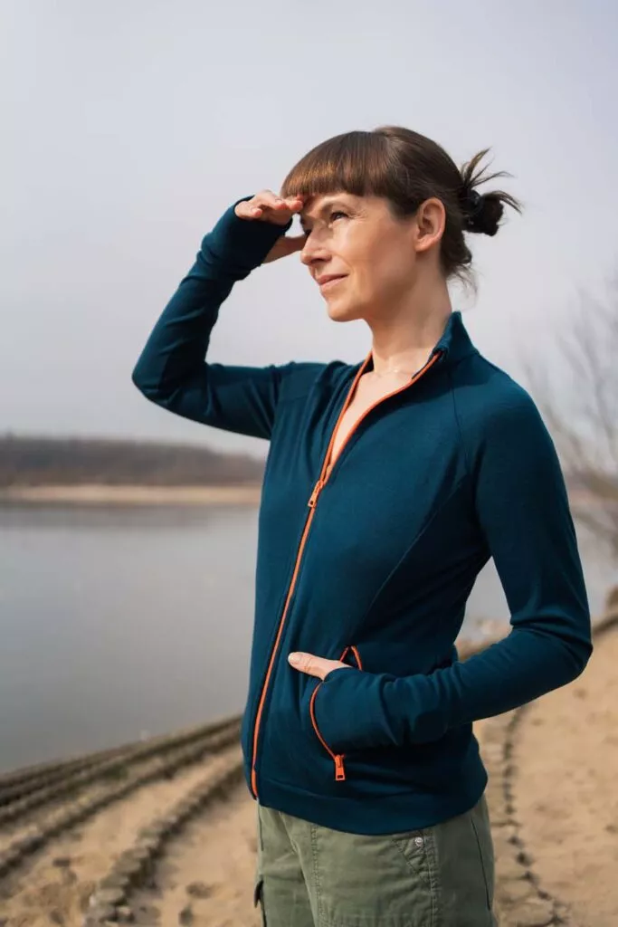 Žena v Merino mikině sportovní dámské - petrol (250 g) z wełny merino 100% stojí venku u vody, stíní si oči a usmívá se.