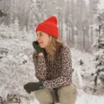 Žena v Merino tričku active se stojáčkem dámské-leopard (135 g) ze 100% vlny merino dřepí ve sněhu a usmívá se.