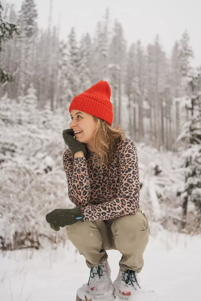 Žena v Merino tričku active se stojáčkem dámské-leopard (135 g) ze 100% vlny merino dřepí ve sněhu a usmívá se.