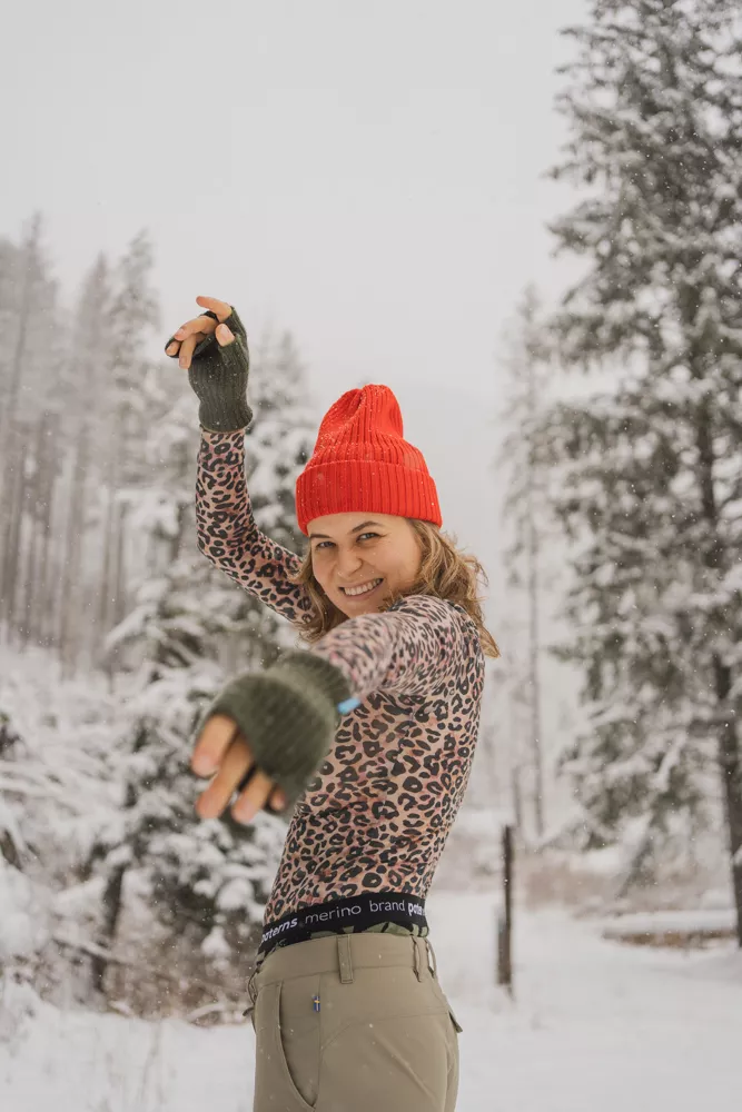 Usměvavá osoba v Merino tričku active se stojáčkem dámské- leopard (135 g) z wełny merino 100% a červené čepici pózuje hravě ve sněhovém lese, v pozadí zasněžené stromy a padající vločky.