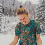 Mladá žena se světle hnědými vlasy v drdolu se směje venku ve sněhovém lese, na sobě má Merino tričko z wełny merino 100%.