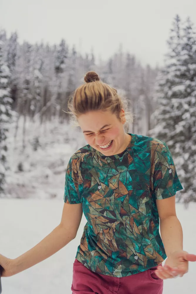 Mladá žena se světle hnědými vlasy v drdolu se směje venku ve sněhovém lese, na sobě má Merino tričko z wełny merino 100%.