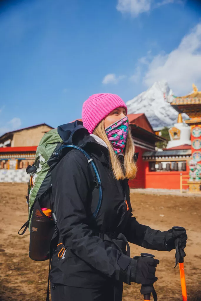 Žena v Merino multifunkčním nákrčníku - pinkpilio (135 g) ze 100% merino vlny, s trekovými holemi, v černém outdooru a růžové čepici, před barevnými budovami a zasněženými horami.