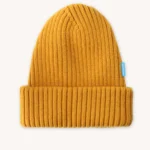 Hořčicově žlutá žebrovaná pletená čepice s ohrnutou manžetou a malou modrou visačkou na pravé straně, zobrazená na hladkém světlém pozadí.