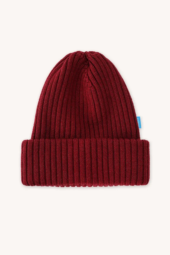 Žebrovaná pletená čepice s ohrnutou manžetou a malou modrou visačkou na boku, zobrazená na hladkém světlém pozadí.