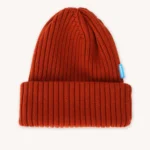 Červená pletená čepice s širokou žebrovanou ohrnovací manžetou a malou modrou visačkou na boku, zobrazená na bílém pozadí.