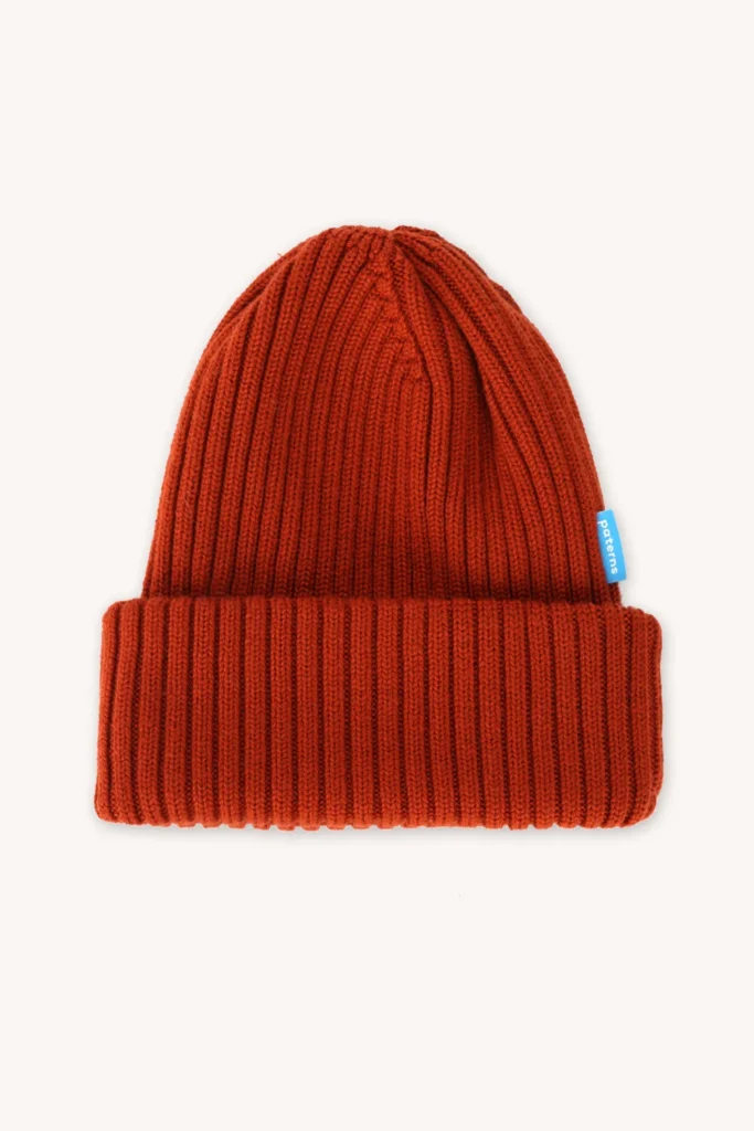 Červená pletená čepice s širokou žebrovanou ohrnovací manžetou a malou modrou visačkou na boku, zobrazená na bílém pozadí.