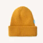 Hořčicově žlutá pletená čepice s ohrnutou manžetou, s malou modrou visačkou na boku, zobrazená na světlém pozadí.