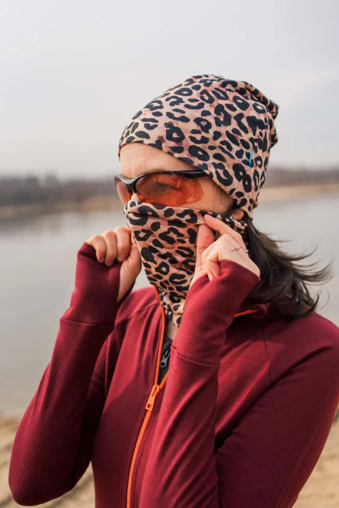 Osoba v klobouku s leopardím vzorem a odpovídajícím krytem obličeje si oběma rukama upravuje masku. Na sobě má sluneční brýle a bordó bundu, stojí venku u vody v zamračeném dni.