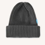 Tmavě šedá pletená čepice s žebrovaným lemem a malou modrou visačkou na boku, zobrazená na světle béžovém pozadí.