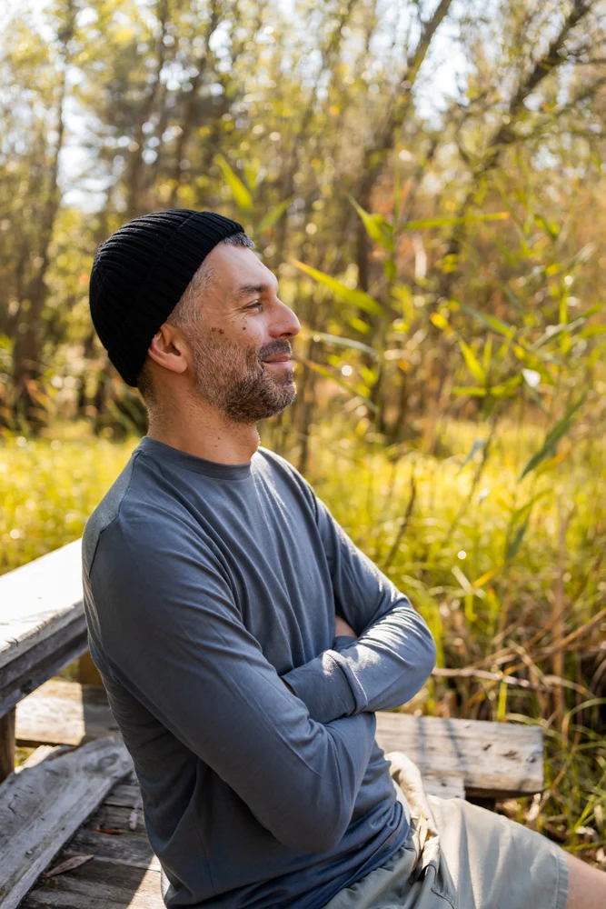 Muž s plnovousem v černé pletené čepici a šedé košili s dlouhým rukávem sedí venku na dřevěné lavičce, usmívá se a užívá si slunečného prostředí plného přírody.