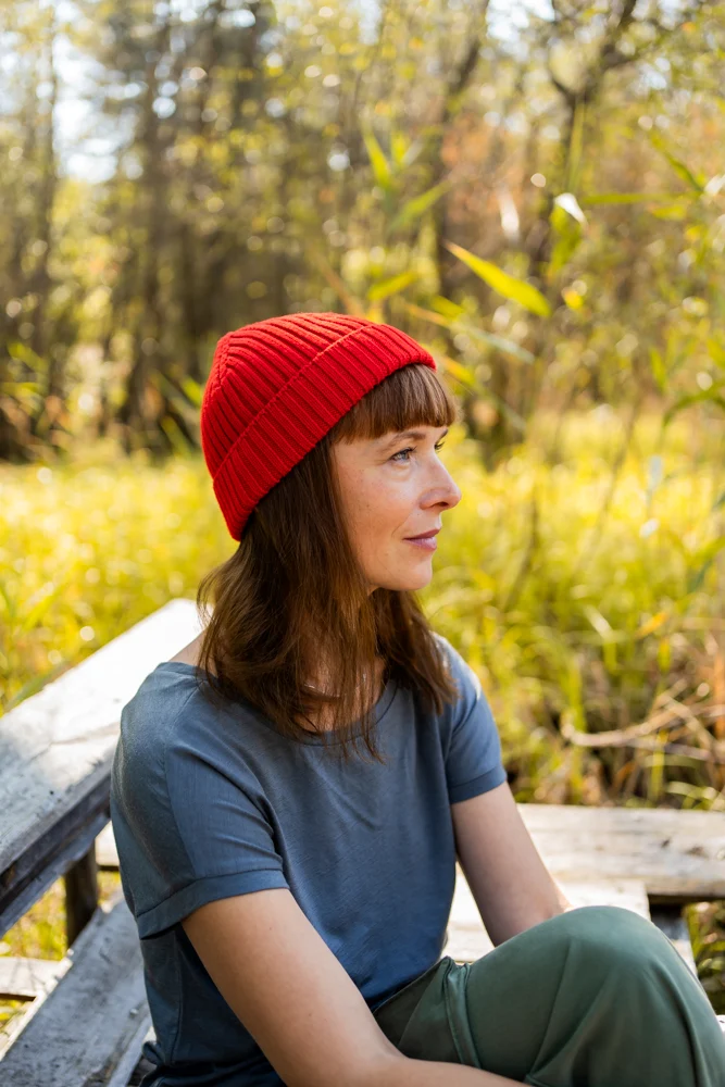 Hnědovlasá žena v červené pletené čepici a modré košili sedí na dřevěné lavičce venku, obklopená vysokou trávou a stromy v přírodním prostředí ozářeném sluncem.