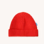 Jasně červená pletená čepice s ohrnutým okrajem a malou modrou visačkou na boku, zobrazená na hladkém bílém pozadí.