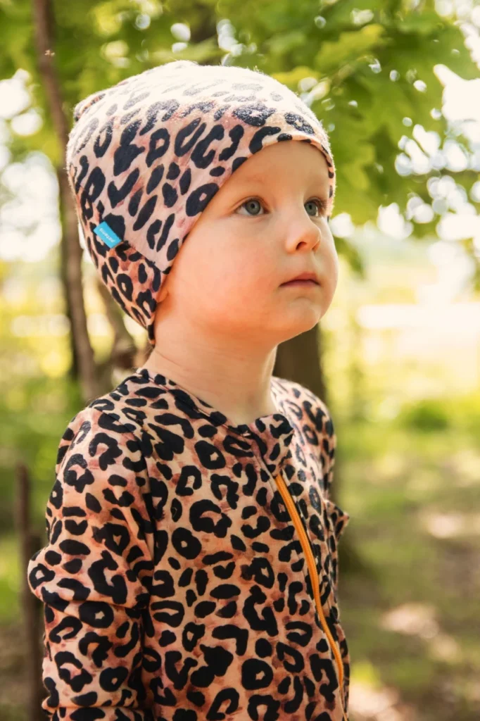 Venku stojí malé dítě ve stejném oblečení a čepici s leopardím vzorem a dívá se mírně vzhůru. Sluneční světlo proniká zelenými listy v pozadí a vytváří tak teplé, přirozené prostředí.