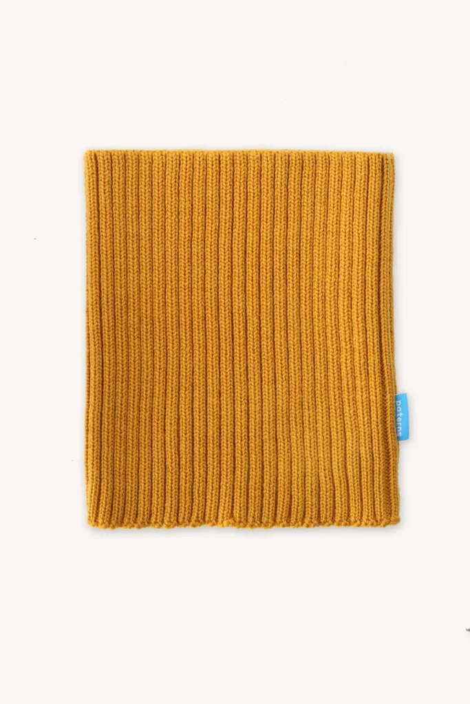 Čtvercová hořčicově žlutá žebrovaná pletenina s malou modrou visačkou na pravém okraji, položená na světlém podkladu.