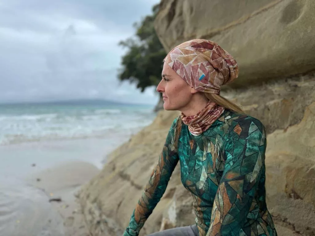 Žena v Merino multifunkčním nákrčníku - ginger gorpcore (135 g) ze 100% merino vlny a s dlouhým rukávem sedí na kamenité pláži, zamyšleně hledí na oceán pod zataženou oblohou.