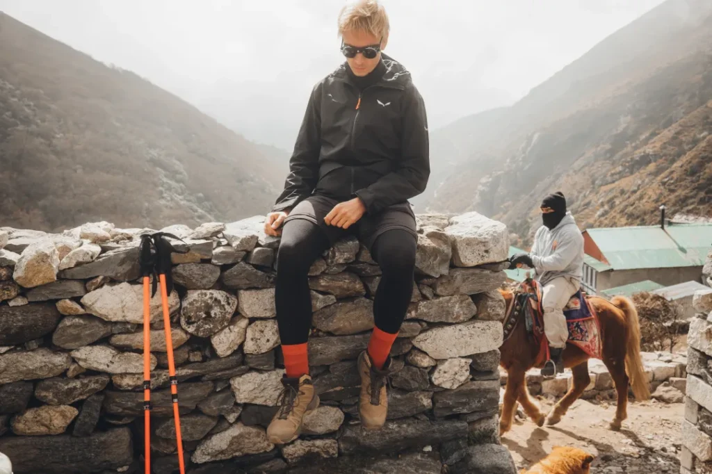 Osoba v turistické výstroji sedí na kamenné zídce v horské krajině a vedle ní se opírají trekingové hole. V pozadí jede jiná osoba na koni po cestě. Po obou stranách se zvedají mlhavé hory.