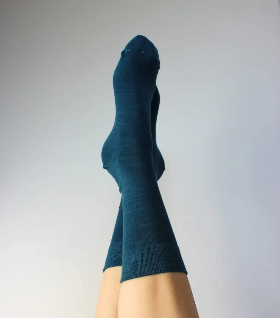 Pár zkřížených nohou v čajových ponožkách je vyobrazen na hladkém světle šedém pozadí.