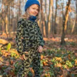 Dítě v topu s dlouhým rukávem z wełny merino 100% - olivové trojúhelníky (190 g), modré čepici a šále stojí sebevědomě mezi slunečnými podzimními listy v lese.
