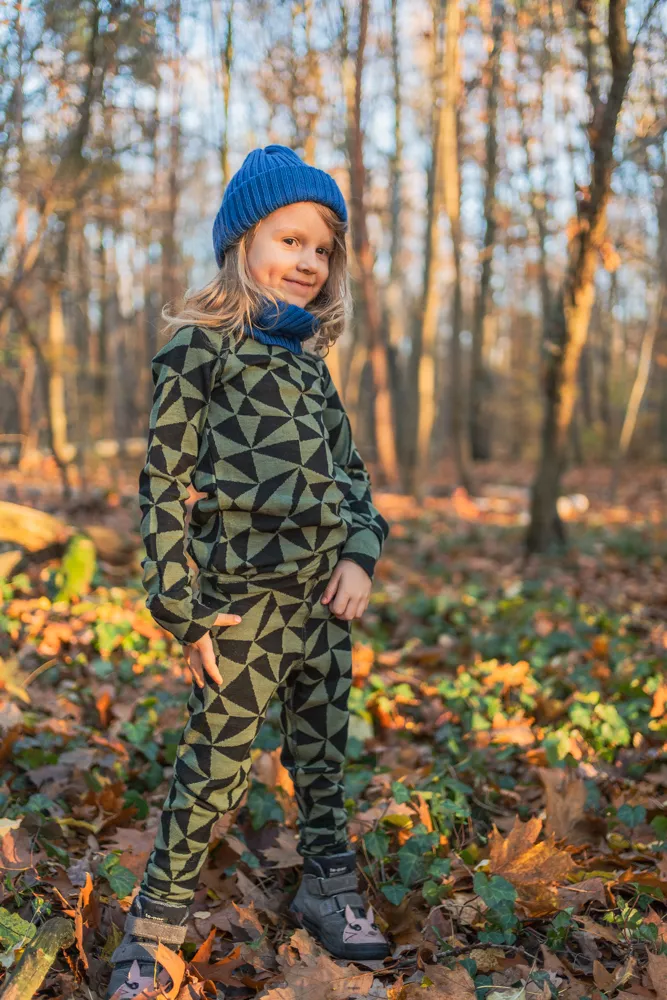 Dítě v topu s dlouhým rukávem z wełny merino 100% - olivové trojúhelníky (190 g), modré čepici a šále stojí sebevědomě mezi slunečnými podzimními listy v lese.