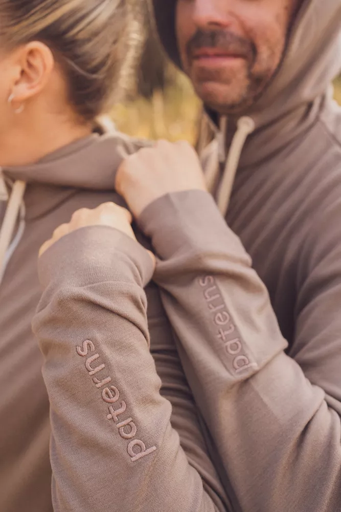Dva lidé v ladících světle hnědých mikinách s nápisem patērns na rukávech z wełny merino 100%. Stojí blízko u sebe venku, částečně otočení zády ke kameni.