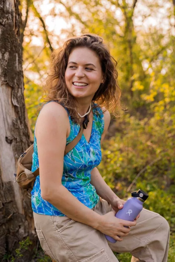 Žena s kudrnatými vlasy sedí venku u stromu a usmívá se. Má modrý vzorovaný top bez rukávů z wełny merino 100%.