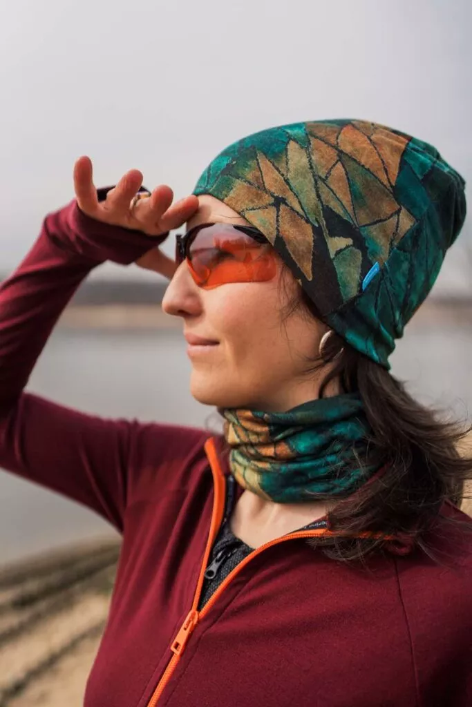 Žena v barevné čepici a nákrčníku z wełny merino 100 %, červené bundě a oranžových brýlích stojí u vody, kryje si oči.
