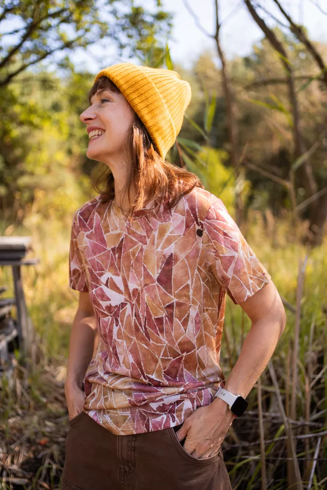 Usměvavá žena v žluté čepici z wełny merino 100 %, vzorovaném triku a chytrých hodinkách stojí venku mezi stromy za slunného dne.