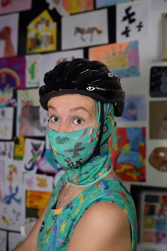 Osoba w kasku rowerowym i kolorowej osłonie twarzy z wełny merino 100% (merino vlna 100%), na tle ściany z rysunkami dzieci.